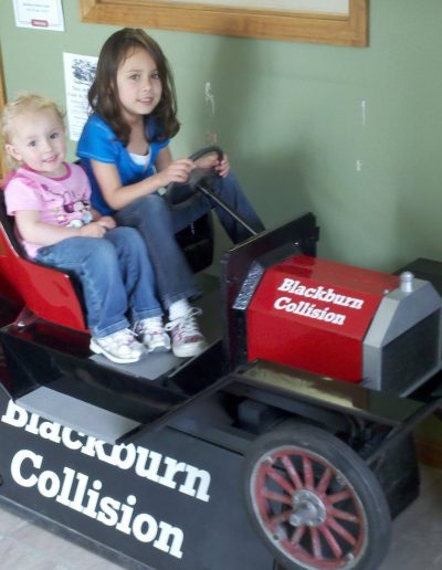 Two small girls posing on a toy car at Blackburn Collision Center
